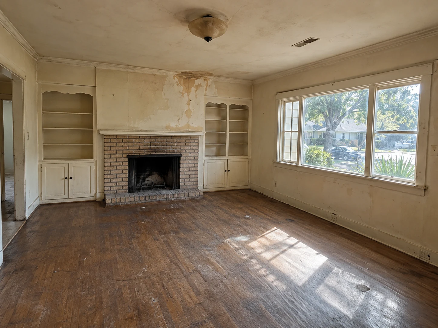 Before concept: an empty living room with worn wood flooring, peeling walls, a brick fireplace, built-in shelves and a large street-facing window
